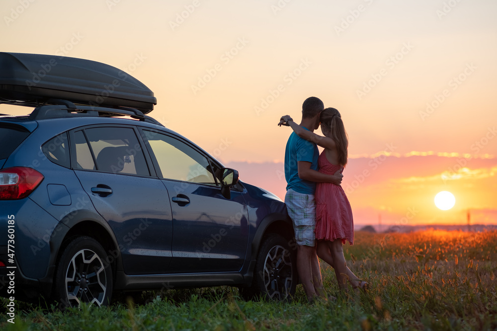 Wall mural happy couple relaxing beside their suv car during honeymoon road trip at sunset. young man and woman