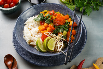 A blue bowl with red thai vegetable curry and basmati rice, fresh lime, cilantro, spinach, chilli peppers and cherry tomatoes
