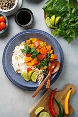 A blue bowl with red thai vegetable curry and basmati rice, fresh lime, cilantro, spinach, chilli peppers and cherry tomatoes, top view