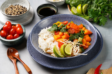 A blue bowl with red thai vegetable curry and basmati rice, fresh lime, cilantro, spinach, chilli peppers and cherry tomatoes