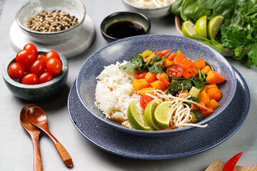 A blue bowl with red thai vegetable curry and basmati rice, fresh lime, cilantro, spinach, chilli peppers and cherry tomatoes