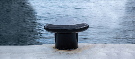 Black bollard on harbor pier, sea water background, space. Metal post anchored on concrete floor © Rawf8
