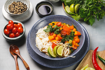 A blue bowl with red thai vegetable curry and basmati rice, fresh lime, cilantro, spinach, chilli peppers and cherry tomatoes