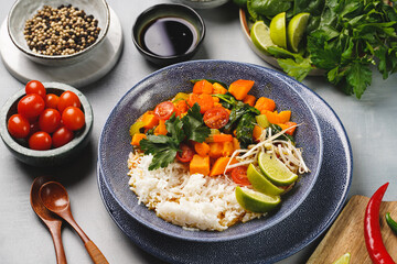 A blue bowl with red thai vegetable curry and basmati rice, fresh lime, cilantro, spinach, chilli peppers and cherry tomatoes