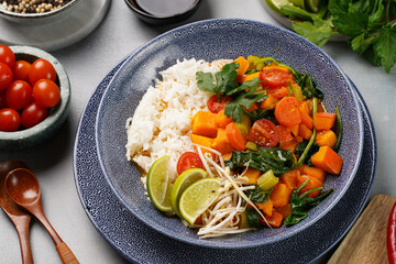 A blue bowl with red thai vegetable curry and basmati rice, fresh lime, cilantro, spinach, chilli peppers and cherry tomatoes