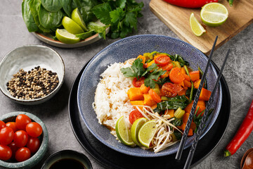 A blue bowl with red thai vegetable curry and basmati rice, fresh lime, cilantro, spinach, chilli peppers and cherry tomatoes