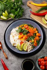 A blue bowl with red thai vegetable curry and basmati rice, fresh lime, cilantro, spinach, chilli peppers and cherry tomatoes, top view