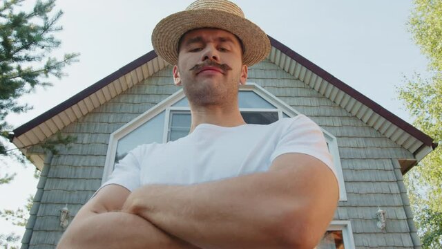 Portrait of a young happy stylish millennial hipster guy with moustache wearing straw hat and on a background of his summer country house courtyard. Modern generation people lifestyle