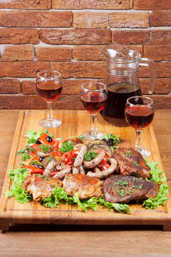 meat and wine on the wooden table. three glasses and carafe. grilled chicken, pork, sausages and vegetables on the board