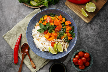 A blue bowl with red thai vegetable curry and basmati rice, fresh lime, cilantro, spinach, chilli peppers and cherry tomatoes, top view