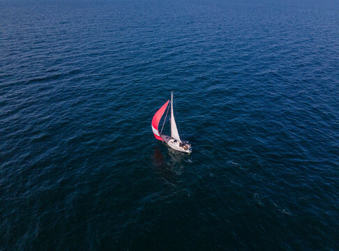 Aerial view of a sailboat with a red spinnaker sail hoisted on a calm and sunny day in the baltic sea, Germany.