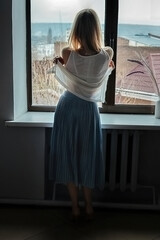 A slender beautiful girl takes off her clothes at home near the window. she has her back turned to us. The cityscape is visible in the window. People at home on vacation after work