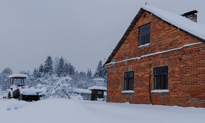 an old red brick house in countryside in winter. in background snowy forest and abandoned little tractor