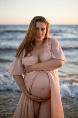 Young pregnant woman with a beautiful sea view on the background. Happy and calm pregnant woman with long hair and pink dress standig on the beach. Romantic view, ocean, sunset, maternity.