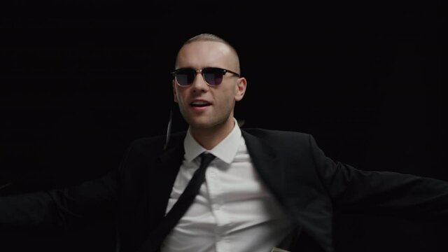 Young happy caucasian businessman in business suit and sunglasses dancing among falling dollar bills in slow motion isolated on black background. Hit a big jackpot. Winner and Successful investor