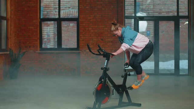 Caucasian woman coach spinning on stationary bike in foggy gym. Athletic girl performing aerobic riding training exercises on cycling bike. Weight Loss Cardio Healthy Lifestyle. Modern sport indoors