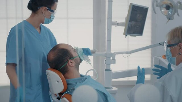 Dental assistant using oxygen mask to do anesthesia on patient before starting surgical procedure at oral care clinic. Nurse giving anesthesic to man, performing stomatological surgery.