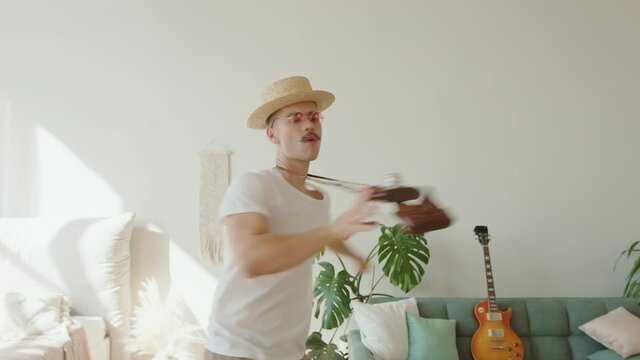 Young fashion creative hipster wearing hat, pink sunglasses guy dancing and rotating around with his old retro fim camera with strap his neck in contemporary inerior. Millennial generation lifestyle