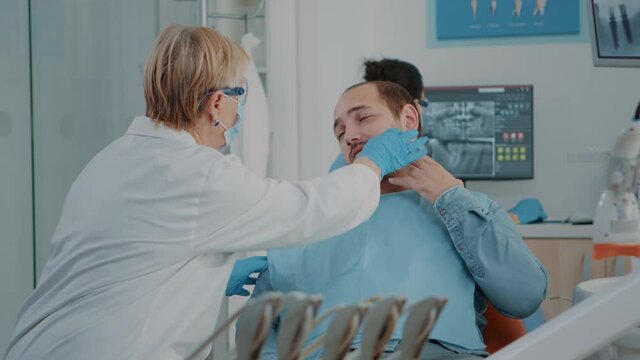 Senior dentist examining patient with serious toothache, getting ready to do stomatological consultation with dental tools in cabinet. Medic treating man in pain with oral care problems.