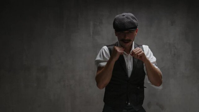 Young dangerous gangster man in vintage suit, cap, rolls up his sleeves, prepares to fight, waving his fists, make punch, uppercut, provoking, stands in fighting stance in front of concrete wall