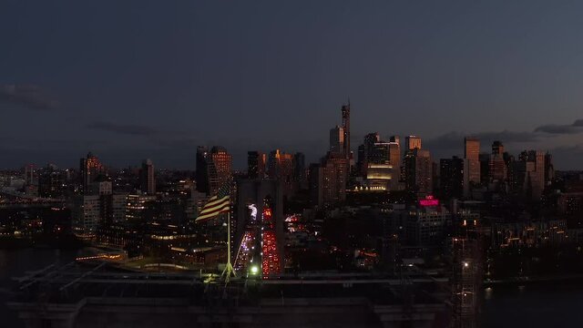 Forwards fly above American flag on top of Brooklyn Bridge. Heavy traffic in evening city. Panoramic view of high rise buildings at dusk. Brooklyn, New York City, USA