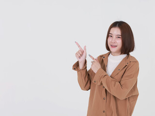 Portrait closeup studio cutout shot of Asian young beautiful short hair female model in brown coat jacket standing smiling look at camera pointing finger up on blank copy space on white background