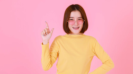 Portrait studio cutout shot Asian young pretty short hair female stylish fashionable model in yellow long sleeve shirt standing look at camera holding adjusting fashion sunglasses on pink background