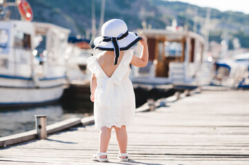 Back view of girl in light dress with hat standing beside docked boats