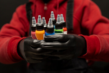 Male tattoo artist holding bottles of tattoo ink of various colors. Selective focus on the jars,...