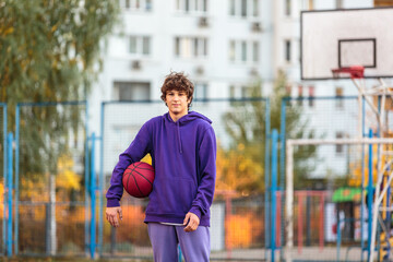 Cute teenager in violet hoodie playing basketball. Young boy with ball learning dribble and shooting on the city court. Hobby for kids, active lifestyle