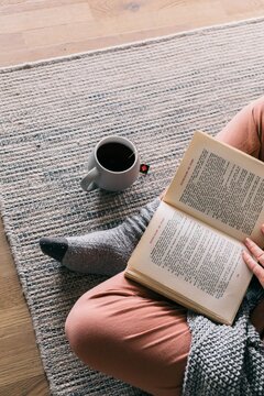 Cropped image of a person wearing socks reading a book and sitting on rug beside cup of tea