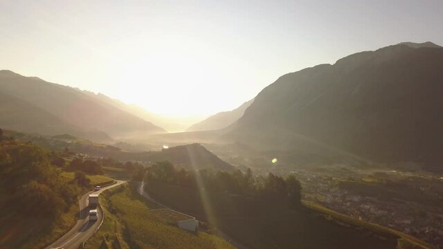 Drone shot in the early morning with the sun rising over a beautiful mountainous valley with some traffic on the road in Lavaux Oron, Switzerland in 4k.