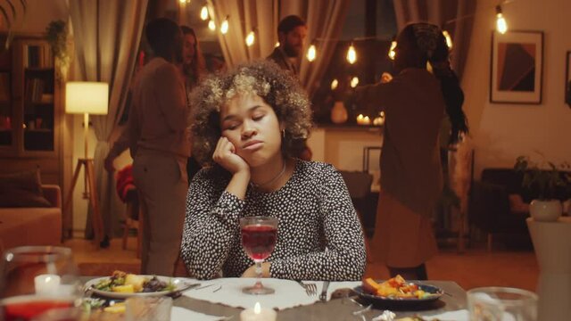 Slow motion of sad young Black woman sitting at dining table during birthday party, friends dancing behind her