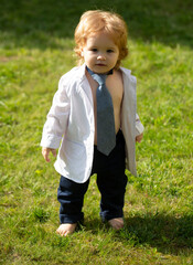 Baby child on the green grass in summer park. Funny little man in suit, jacket and necktie.