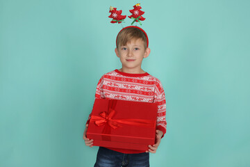 a handsome blond boy in a Christmas tree-shaped headband in a Christmas sweater holds a red gift box in his hands