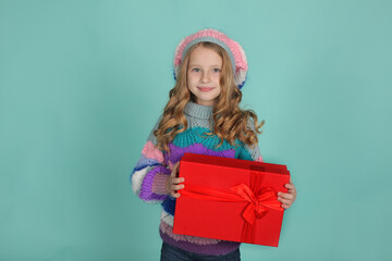 a beautiful happy blonde girl in a colored knitted sweater and hat in her hands a red gift box on a blue background
