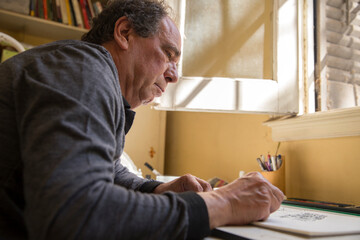 Fototapeta premium Concentrated Senior man enjoying while drawing a pastel sketch at home studio while daylight gets into a window