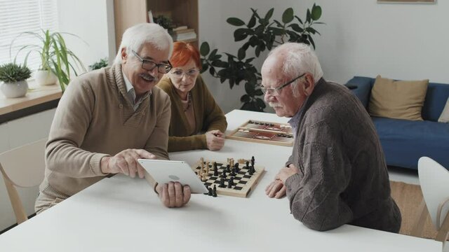 Medium shot of three modern seniors using digital tablet and talking while sitting at table with chess and backgammon games on it