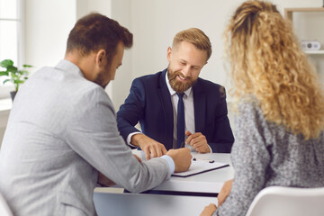 Young couple buying or renting house. Smiling real estate agent gives clients papers to sign. Happy buyers or tenants sitting together at desk in realtor's office and putting signatures on agreement