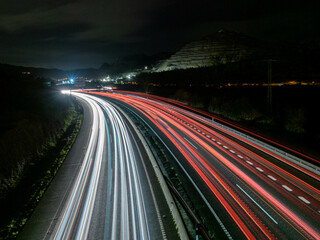 Trazos de luz dejados por el tráfico al circular por la noche por una autopista a su paso por un desfiladero, con las luces de una ciudad al fondo.