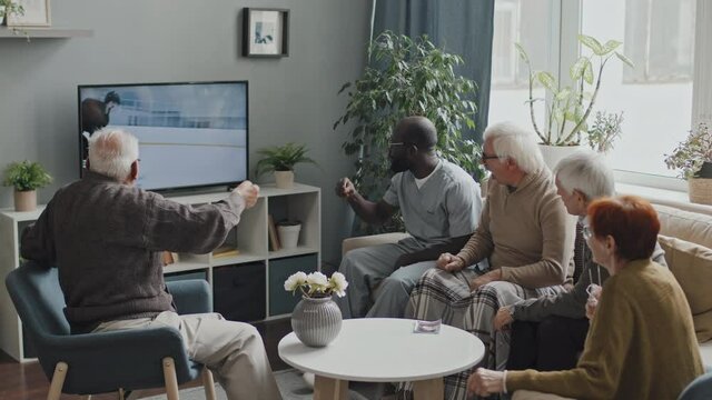 Four Caucasian seniors and African-American male nurse or social worker watching exciting hockey game on TV together, sitting on sofa in cozy living room at nursing home