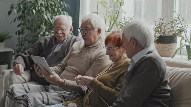 Medium shot of four seniors having discussion while using digital tablet, sitting together on sofa at nursing home