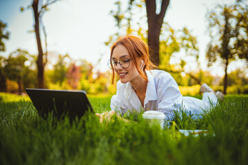 beautiful smiling girl student uses laptop pc while lying on the grass outdoors. Education concept