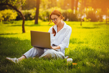 Redhead freelancer girl glasses works with laptop, outdoors while sitting on the grass. Freelance 
