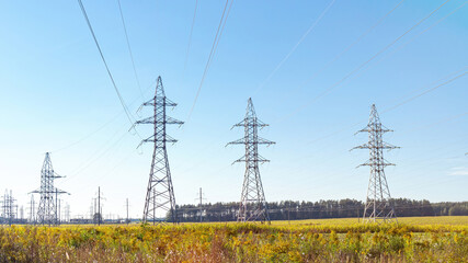 High voltage towers with blue sky background. Overhead power lines on the background of forest and green field. Electricity transmission through power lines. Green energy.