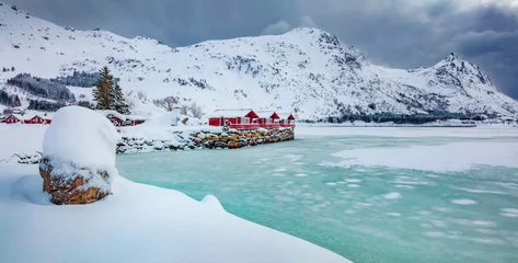 Fotobehang Life over polar circle. Traditional Norwegian red wooden houses on the shore of frozen fjord. Dramatic winter view of Kongsjordpollen fjord, Lofoten islands, Vestvagoy, Norway, Europe. © Andrew Mayovskyy