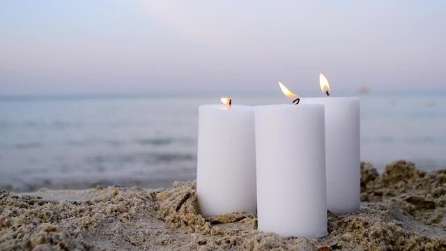 Three large white paraffin candles burn on sand beach background sea waves. Decorative Burning candles on sea coast close up. Candlelight, candle flame. Romantic mood backdrop. Concept date relaxation
