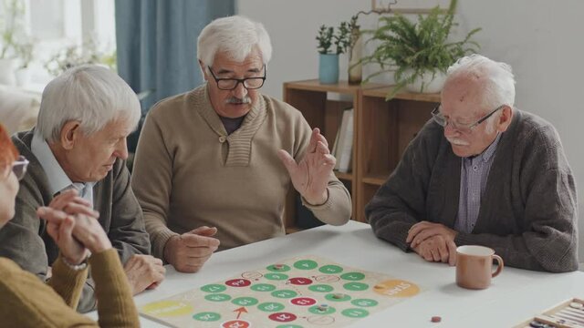 Medium shot of group of happy senior people playing table game together in living room of nursing home