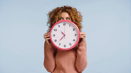tricky woman obscuring face with round clock while looking aside isolated on blue