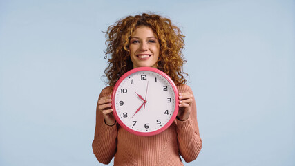 cheerful redhead woman holding white round clock while looking at camera isolated on blue